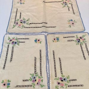 Könnte beinhalten: Drei rechteckige Platzsets aus cremefarbenem Stoff mit gestickten Blumensträußen in Rosa, Blau und Gelb sowie grünen Blättern. Jedes Platzset ist mit schwarzen, leiterartigen Stickereien und einem hellblauen Häkelrand versehen.