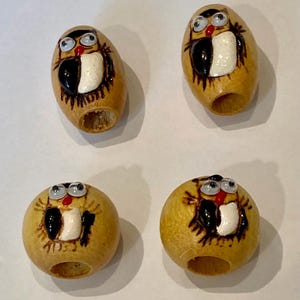 May include: Four light brown wooden beads, each featuring a hand-painted owl design with black and white details, a small red beak, and two attached googly eyes. The beads have a central hole for threading and vary in shape between oval and round.