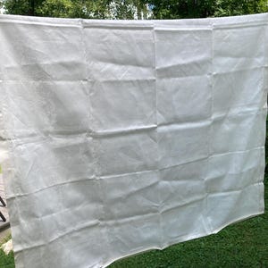 May include: A large, rectangular white fabric sheet featuring a subtle, repeating tonal damask pattern. The material is lightweight and semi-translucent, displaying visible fold lines across its surface. The edges are finished with a simple hem.