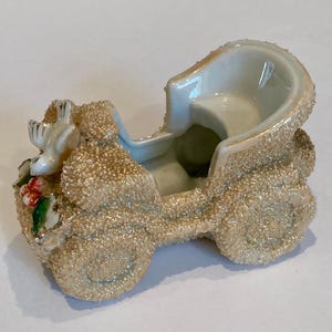 May include: A ceramic figurine shaped like a vintage carriage with a textured, sandy beige finish. The front features a small white bird perched near green leaves and red berries. The interior of the carriage is smooth, glossy, and white.