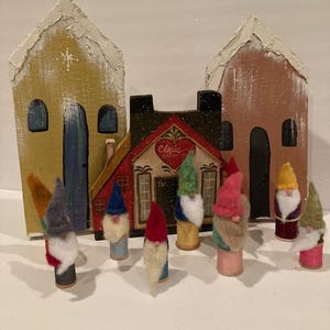 May include: A miniature village scene with two houses, a red building with a heart and the word "Claus" on it, and eight felt gnomes with different colored hats and beards.