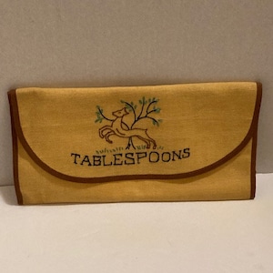 Op de afbeelding: Een rechthoekig mosterdgeel stoffen tasje met een bruine bies en een klepsluiting. De voorzijde is voorzien van een geborduurd ontwerp van een hert onder een boom met groene bladeren en het woord TABLESPOONS gestikt in donkerblauw garen.
