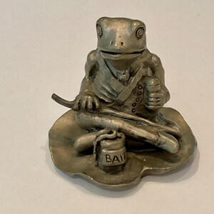 Op de afbeelding: Een metalen zilverkleurig beeldje van een kikker die op een waterlelieblad zit. De kikker draagt een vest, houdt een hengel vast en voor de gekruiste benen staat op het blad een klein cilindervormig potje met het opschrift "BAIT".