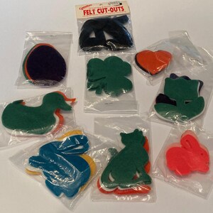 May include: A collection of assorted felt cut-outs in various shapes including a duck, rabbit, cat, clover, heart, and tulip, packaged in individual clear plastic bags. The top package is labeled Felt Cut-Outs for Quick N Easy Craft & Clothing Decorating.