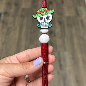 May include: A red glitter pen with a Day of the Dead skull topper wearing a green and red sombrero. The pen has a silver tip and is held in a hand with a light purple painted nail.