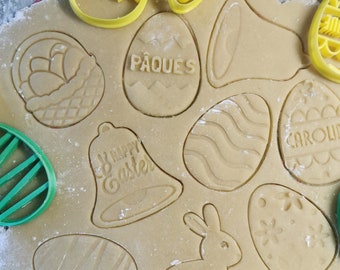 May include: A close-up of a baking sheet with various cookie cutters and dough cutouts. The cutters include a bunny, a bell, an egg, and a basket. The dough cutouts are decorated with words like "Pâques" and "Happy Easter".