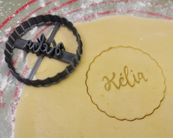 May include: A black plastic cookie cutter with the name "KÉLIA" cut out. The cutter is on top of a sheet of yellow cookie dough with the name "KÉLIA" pressed into it.