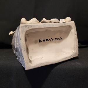 Op de afbeelding: Een wit keramisch beeld van een hoofd met een zwarte inscriptie op de onderkant die "AMASSIDA" luidt.
