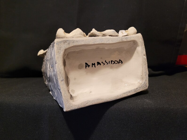 Op de afbeelding: Een wit keramisch beeld van een hoofd met een zwarte inscriptie op de onderkant die "AMASSIDA" luidt.