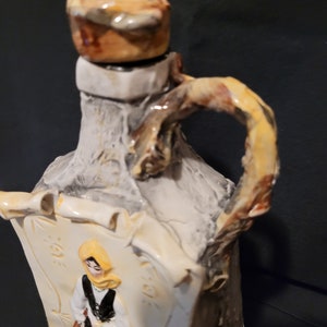 Op de afbeelding: Een keramische fles met een rustieke, getextureerde afwerking. De fles heeft een kurkstop en een handvat in de vorm van een tak. De fles heeft een geschilderd beeld van een vrouw in traditionele kleding.