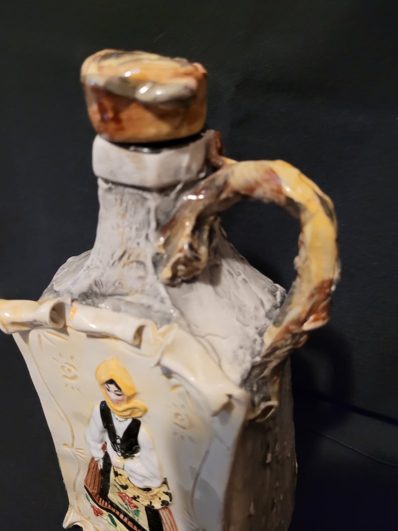 Op de afbeelding: Een keramische fles met een rustieke, getextureerde afwerking. De fles heeft een kurkstop en een handvat in de vorm van een tak. De fles heeft een geschilderd beeld van een vrouw in traditionele kleding.