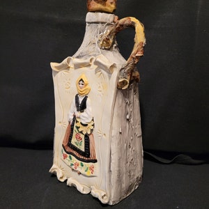 Op de afbeelding: Een keramische fles met een rustieke, hout-achtige afwerking. De fles heeft een reli&euml;fbeeld van een vrouw in traditionele kleding, met een gele hoofddoek, een zwart lijfje en een kleurrijke rok. De fles heeft een handvat in de vorm van een tak.