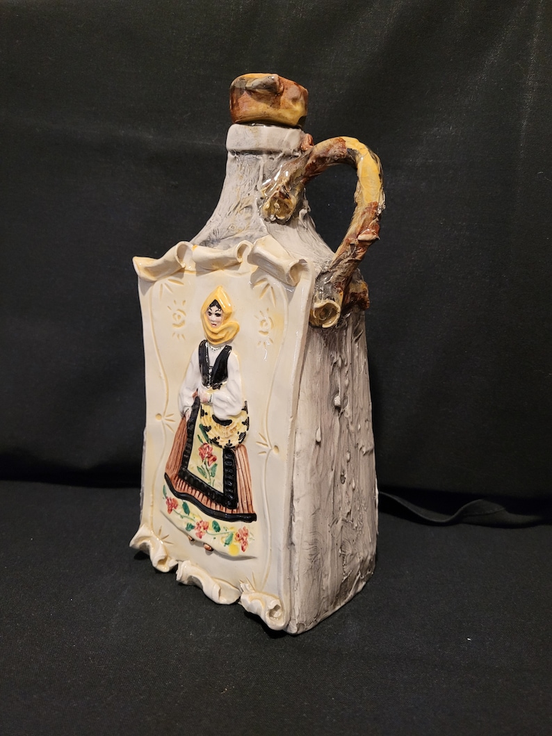 Op de afbeelding: Een keramische fles met een rustieke, hout-achtige afwerking. De fles heeft een reli&euml;fbeeld van een vrouw in traditionele kleding, met een gele hoofddoek, een zwart lijfje en een kleurrijke rok. De fles heeft een handvat in de vorm van een tak.