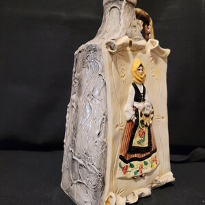 Op de afbeelding: Een keramische fles met een wit en grijs getextureerd oppervlak. De fles heeft een reli&euml;fbeeld van een vrouw in een gele hoofddoek en een bruin en zwart jurkje. De vrouw houdt een boeket bloemen vast.