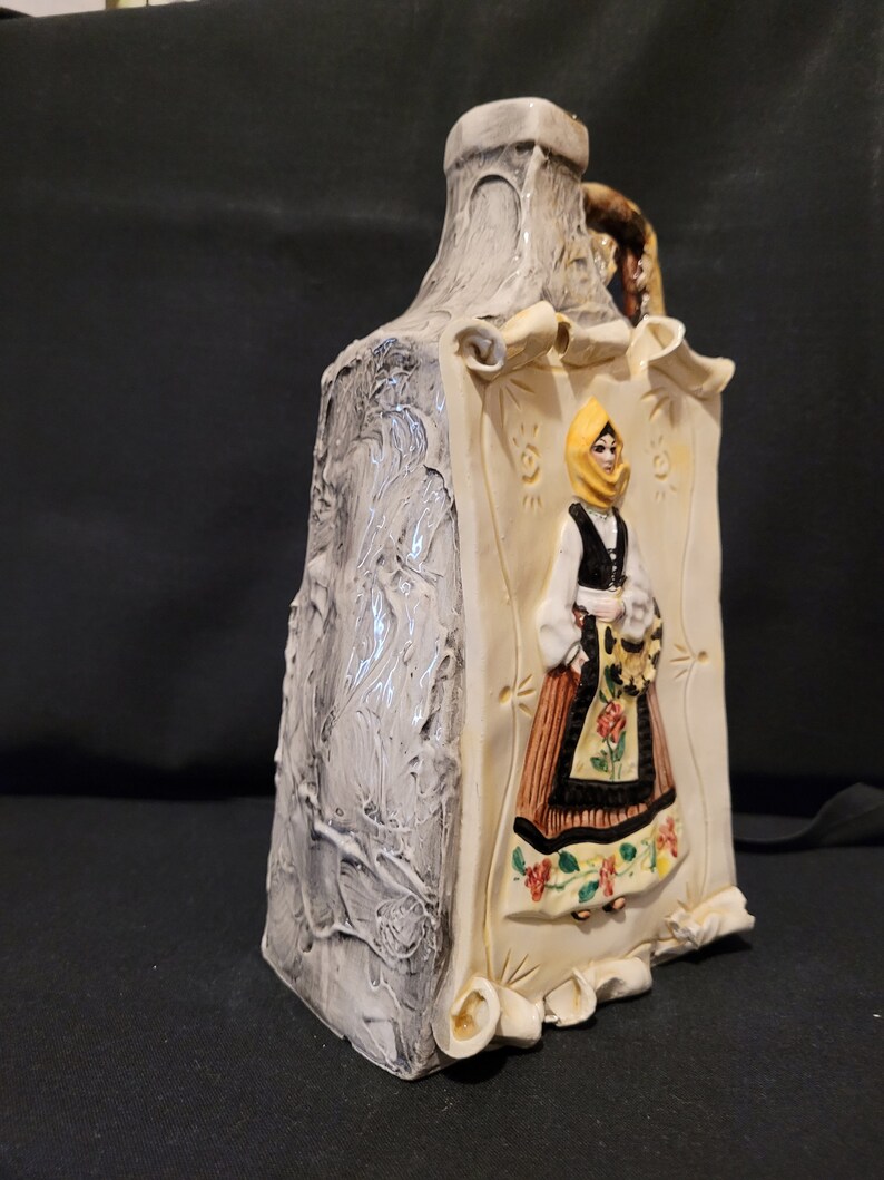 Op de afbeelding: Een keramische fles met een wit en grijs getextureerd oppervlak. De fles heeft een reli&euml;fbeeld van een vrouw in een gele hoofddoek en een bruin en zwart jurkje. De vrouw houdt een boeket bloemen vast.