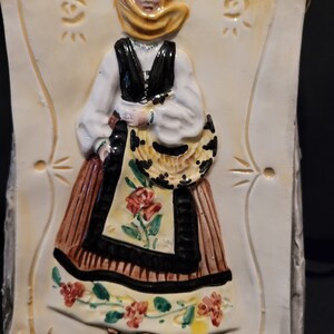 Op de afbeelding: Een keramische wandplaat met een vrouw in een gele hoofddoek en een bruin en zwart jurkje met bloemenaccenten. De vrouw houdt een mandje in haar hand.