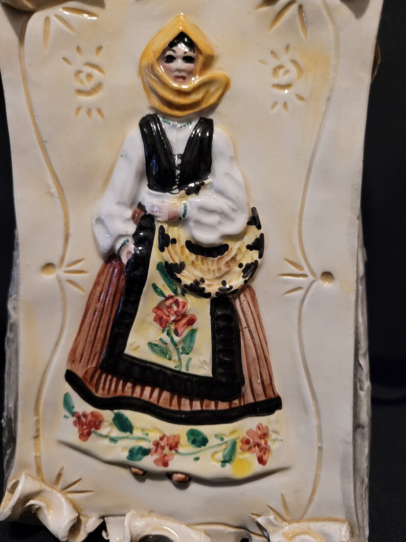 Op de afbeelding: Een keramische wandplaat met een vrouw in een gele hoofddoek en een bruin en zwart jurkje met bloemenaccenten. De vrouw houdt een mandje in haar hand.