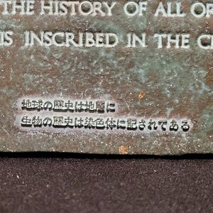 Op de afbeelding: Een close-up van een bronzen plaquette met de tekst "THE HISTORY OF ALL ORGAN IS INSCRIBED IN THE CHRO" en Japanse tekst die vertaalt naar "De geschiedenis van de aarde ligt in de lagen, de geschiedenis van het leven is vastgelegd in de chromosomen."