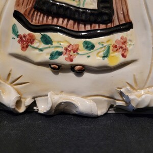 Op de afbeelding: Een keramische tegel met een reli&euml;fbeeld van een vrouw in een bruine en zwarte jurk met een bloemenpatroon. De vrouw staat op een witte basis met een gegolfde rand.