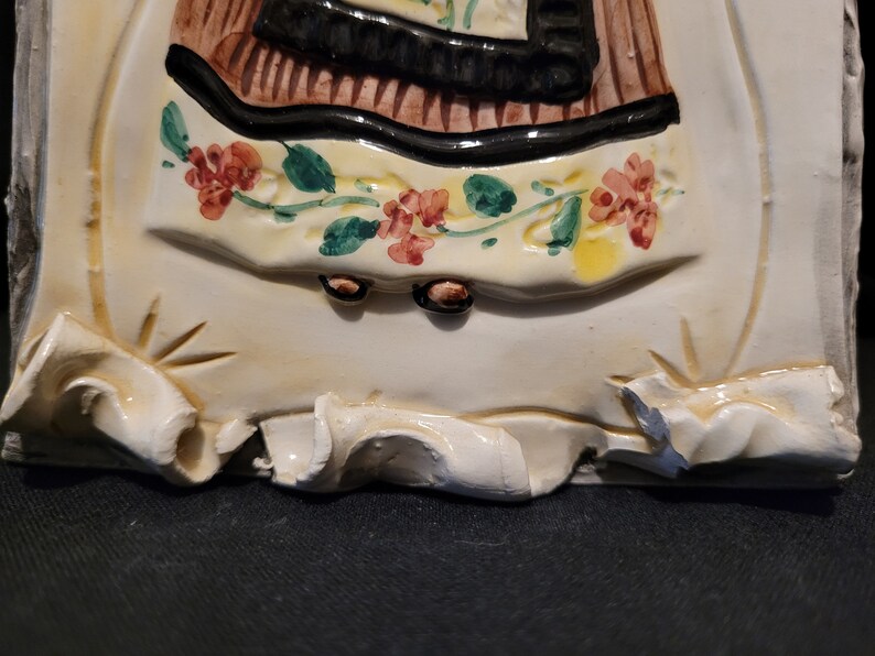 Op de afbeelding: Een keramische tegel met een reli&euml;fbeeld van een vrouw in een bruine en zwarte jurk met een bloemenpatroon. De vrouw staat op een witte basis met een gegolfde rand.