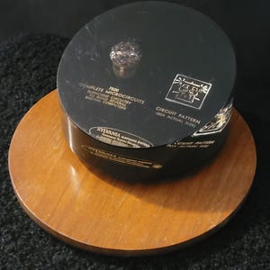 Puede incluir: Un objeto cilíndrico negro con letras doradas y un patrón de circuito, etiquetado como "COMPLETE MICROCIRCUITS" y "CIRCUIT PATTERN (20X ACTUAL SIZE)". Se encuentra sobre una base de madera. El objeto es de Sylvania Electronic Systems.