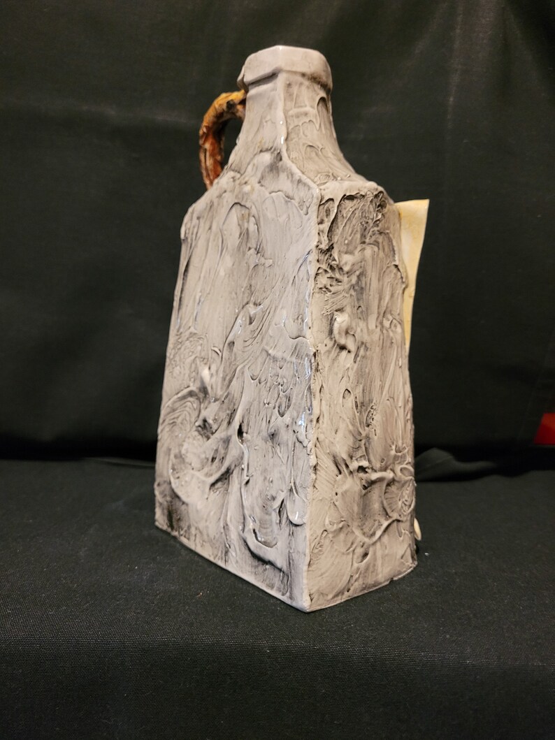 Op de afbeelding: Een witte keramische kan met een getextureerd oppervlak en een bruine handgreep. De kan heeft een uniek, abstract ontwerp.