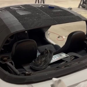 Op de afbeelding: Een witte cabriolet met zichtbaar interieur. De auto heeft zwarte stoelen en een zwart stoffen dak. De auto staat in een garage en heeft een kentekenplaat met de tekst "7EBM".