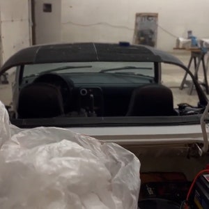 Op de afbeelding: Een witte cabriolet met het dak eraf, waardoor het interieur zichtbaar is. De auto staat in een garage met een plastic zak die de achterbank bedekt.