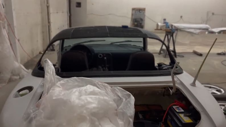 Op de afbeelding: Een witte cabriolet met het dak eraf, waardoor het interieur zichtbaar is. De auto staat in een garage met een plastic zak die de achterbank bedekt.