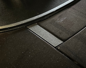 Puede incluir: Primer plano de una superficie negra texturizada con un panel rectangular gris. El panel está integrado en la superficie en ángulo.