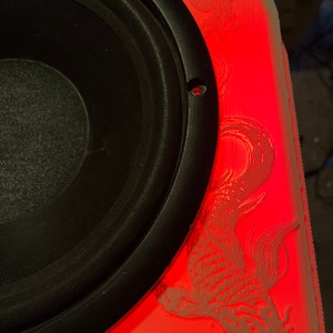 May include: A black speaker with a red illuminated panel. The panel features etched koi fish and dragon designs. The speaker is mounted in a custom enclosure.