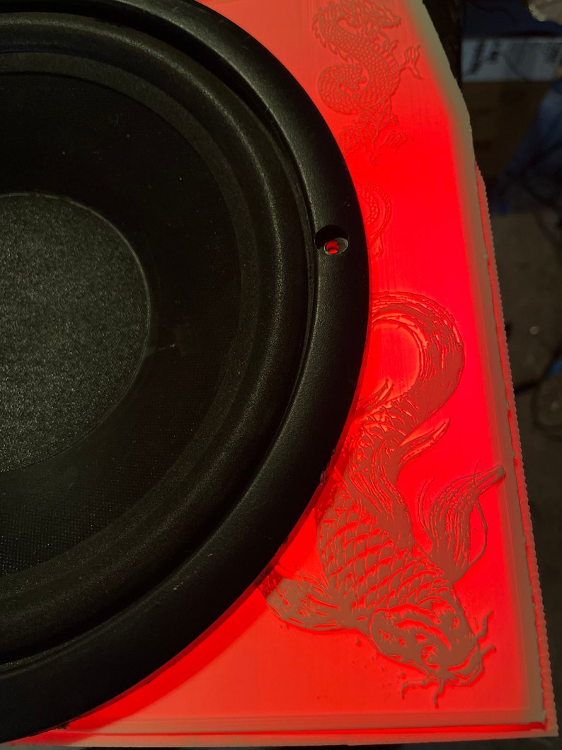 May include: A black speaker with a red illuminated panel. The panel features etched koi fish and dragon designs. The speaker is mounted in a custom enclosure.