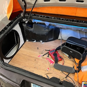 May include: Car trunk interior with a custom audio system installation. Features a subwoofer enclosure, wood-look flooring, and various electronic components, including a multimeter and amplifier. The trunk is open, revealing the installation process.