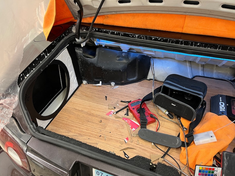 May include: Car trunk interior with a custom audio system installation. Features a subwoofer enclosure, wood-look flooring, and various electronic components, including a multimeter and amplifier. The trunk is open, revealing the installation process.