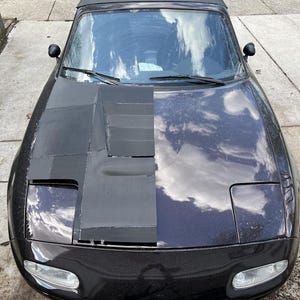 K&ouml;nnte beinhalten: Ein schwarzes Auto mit einer individuellen Motorhaube. Die linke Seite der Motorhaube ist mit schwarzem Material bedeckt, w&auml;hrend die rechte Seite blank ist und die Farbe des Autos zeigt.