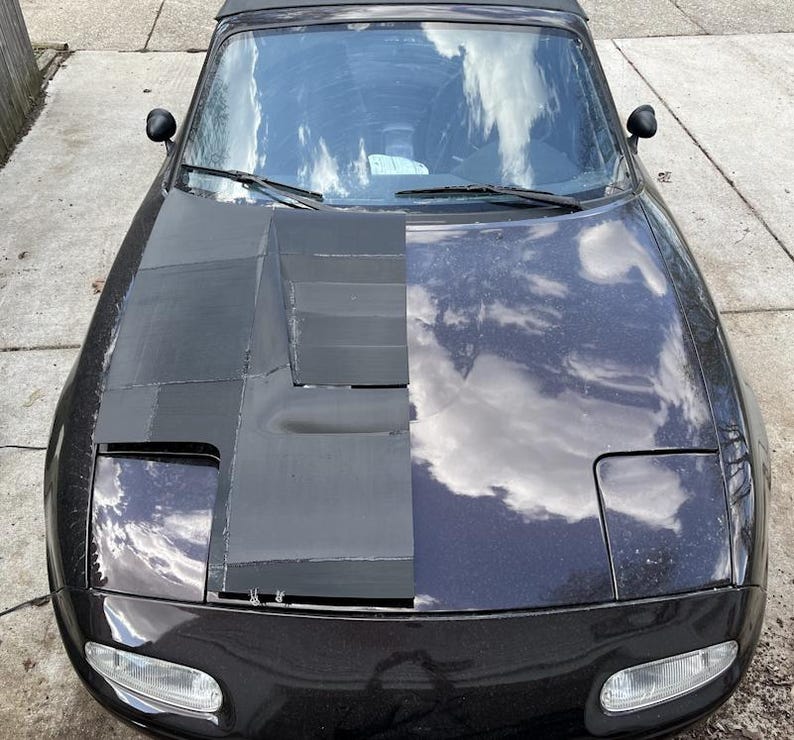 K&ouml;nnte beinhalten: Ein schwarzes Auto mit einer individuellen Motorhaube. Die linke Seite der Motorhaube ist mit schwarzem Material bedeckt, w&auml;hrend die rechte Seite blank ist und die Farbe des Autos zeigt.