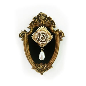 May include: A gold-tone ornate frame with a black velvet backing displays a delicate white and gold floral pendant with a pearl drop.