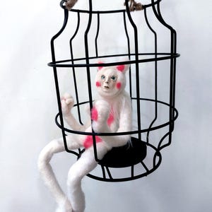 May include: A white needle-felted creature with pink accents on its ears, cheeks, and chest, featuring human-like facial features and long limbs. The creature is seated inside a black wire cage structure suspended by a piece of jute twine.