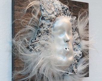 May include: A mixed-media artwork featuring a sculpted face with closed eyes and a slightly open mouth. The face is painted silver and surrounded by white, fluffy material, mounted on a dark wooden square.