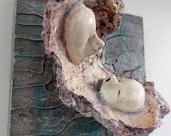 May include: A ceramic wall sculpture featuring two faces, one within the other, on a textured background with shades of brown, blue, and gold.