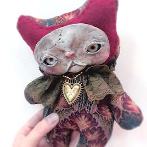 May include: A plush doll with a textured, weathered-looking face, pointed ears, and a body covered in a dark, patterned fabric with deep red and blue tones. The doll features a dark lace collar and a gold-coloured heart pendant on a beaded chain.
