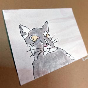 May include: Watercolor illustration of a cat with gray fur, yellow eyes, and a pink nose. The cat is looking up and to the left. The artwork is on a square piece of paper with a light brown background.