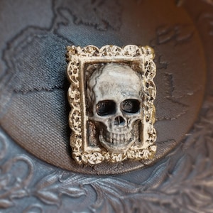 Puede incluir: Un broche rectangular que presenta un cráneo humano tridimensional centrado dentro de un marco dorado texturizado y ornamentado. El cráneo es de color blanco hueso con sombras oscuras en las cuencas de los ojos y la cavidad nasal, con borde de filigrana.