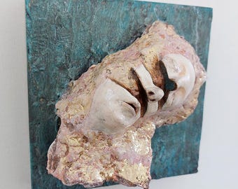May include: A white ceramic sculpture of a face partially embedded in a textured, gold and pink rock. The sculpture is mounted on a square, teal-colored background.