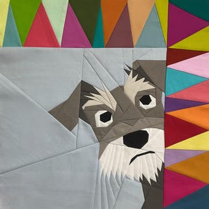 Op de afbeelding: Een stoffen quilt met een geometrisch ontwerp en het gezicht van een hond. De hond is grijs, wit en zwart, met een verdrietige uitdrukking. De quilt heeft een rand van kleurrijke driehoeken in tinten rood, roze, groen en oranje.