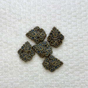 May include: Five decorative ceramic buttons with a fleur-de-lis design. The buttons are a dark bronze colour with blue accents in the detailed scrollwork. The buttons are arranged on a white textured surface.