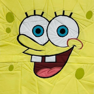 May include: A yellow quilt depicting a cartoon character's face. The character has large white eyes with blue irises, a wide smile with two teeth, and a red nose. The quilt is constructed from geometric shapes.