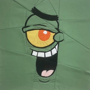 May include: A green fabric art piece featuring a cartoon character's face. The character has a large yellow eye with an orange center, a black eyebrow, and a wide open mouth with white teeth. The background is a patchwork of green fabric.