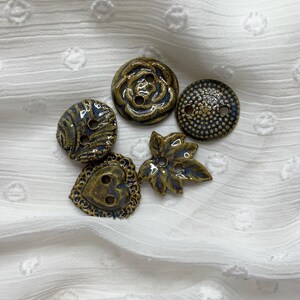 May include: Five ceramic buttons in different shapes, including a rose, a leaf, a heart, and two round designs. The buttons are mainly brown with blue detailing and have two or four holes for sewing.