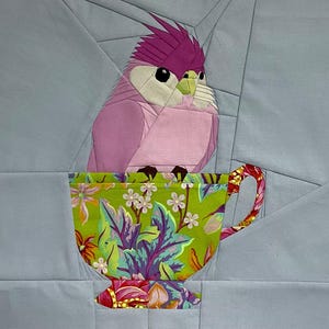 Puede incluir: Una obra de arte textil que presenta un pájaro rosa con una cresta púrpura posado dentro de una taza de té floral. La taza de té tiene un fondo verde con flores y hojas coloridas. El fondo es de color azul claro.